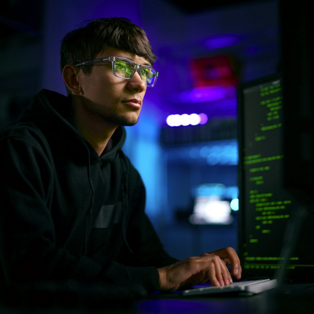 Low Key Lighting Shot Of Male Computer Hacker Sitting In Front Of Screens Breaching Cyber Security